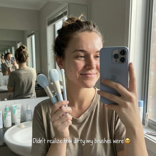 Person taking a mirror selfie with makeup brushes and a phone, in a bathroom setting.